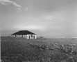 Black and white photograph of a farmhouse on windswept plain, titled "Stone House and Dancing Posts, New Mexico, architectural and landscape photography by Tony Sanders